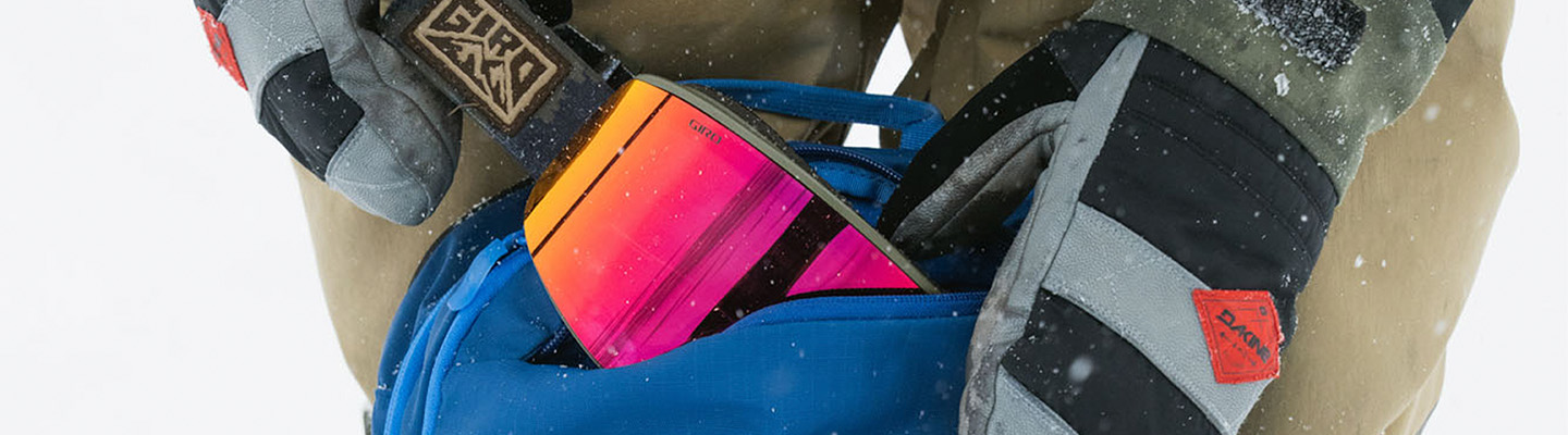 close up image of someone wearing dakine mittens putting an orange and pink pair of giro ski goggles into a blue dakine backpack