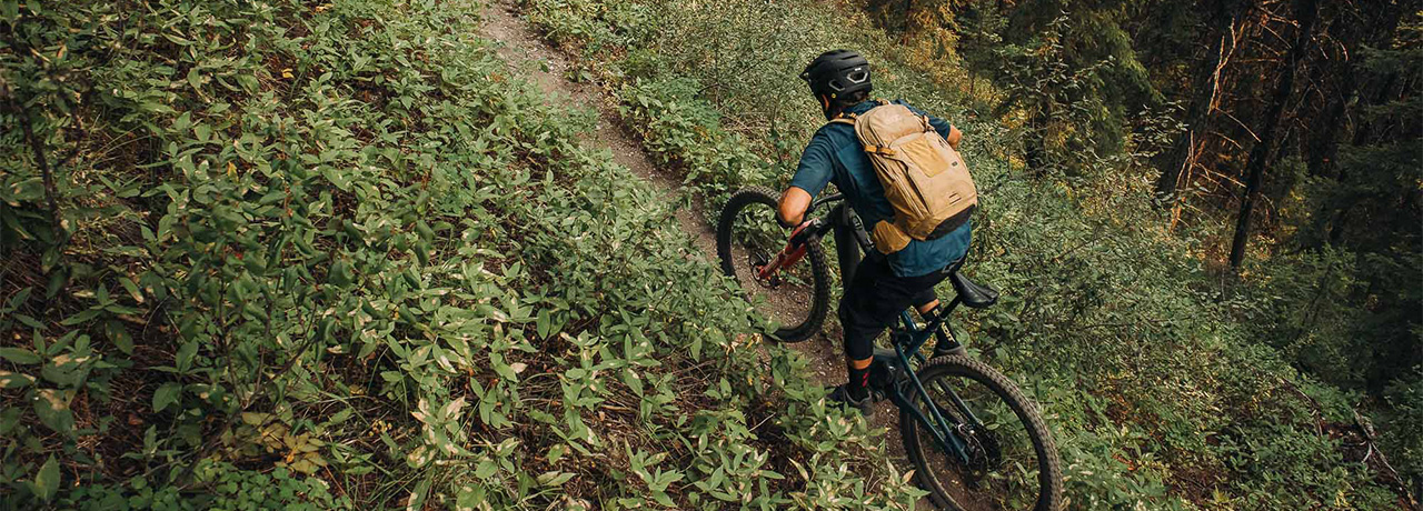 Man wearing a black bike helmet and a tan backpack riding his mountain bike up a single-track path in a forest