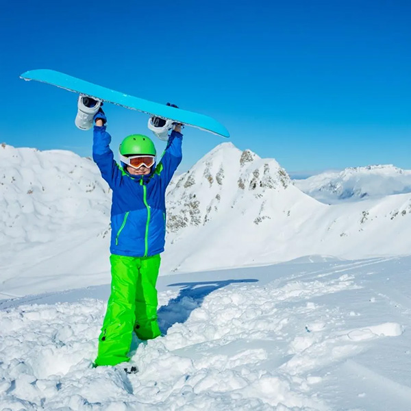 Child wearing blue and green ski gear holding a blue snowboard over their head