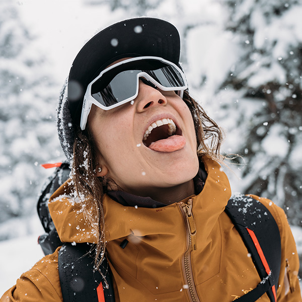 woman in an orange jacket catching snowflakes on her tongue wearing white smith wrap around sunglasses