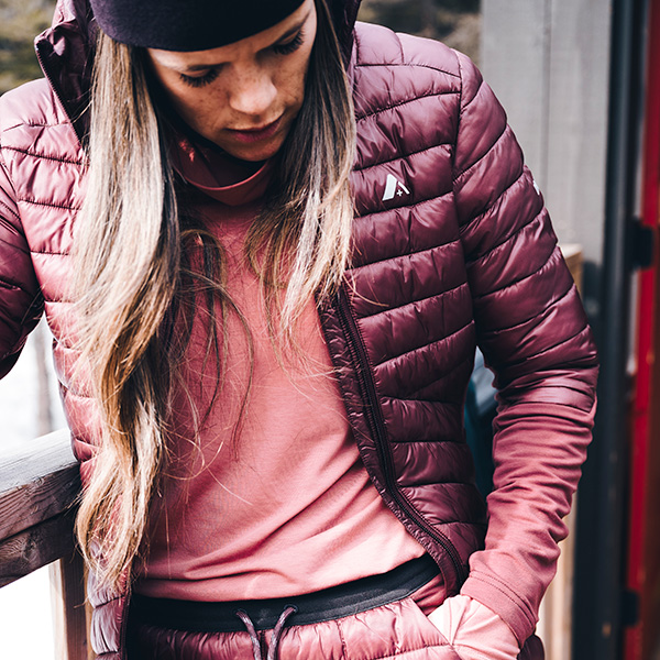 woman wearing dark pink orage base layers, putting on a dark pink orage puffer as a mid layer jacket, also wearing a black beanie hat