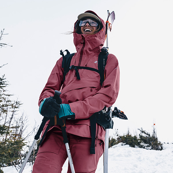 woman carrying skis on her back wearing a dark pink Orage shell jacket and dark pink Orage snow pants