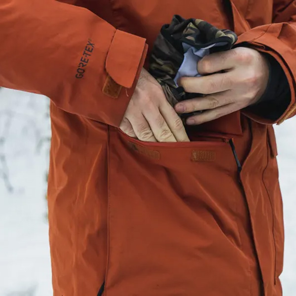 close view of a pocket on an orange shell jacket with someone putting a ski neckie in the pocket
