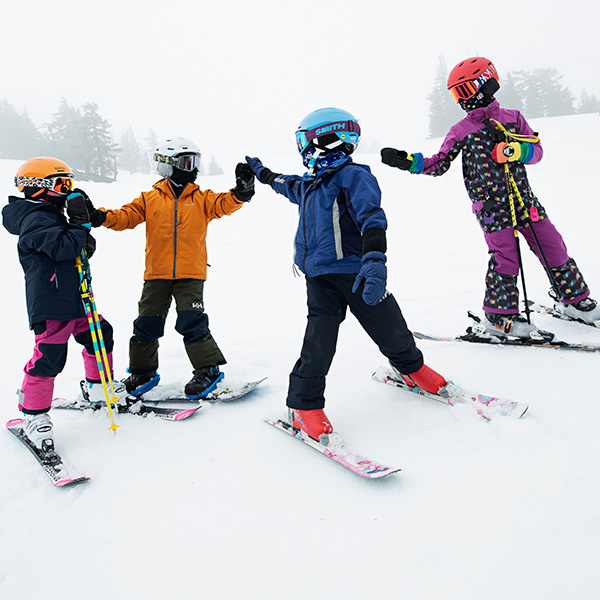 four children in ski gear, ski helmets, and ski goggles high fiving standing on a ski trail