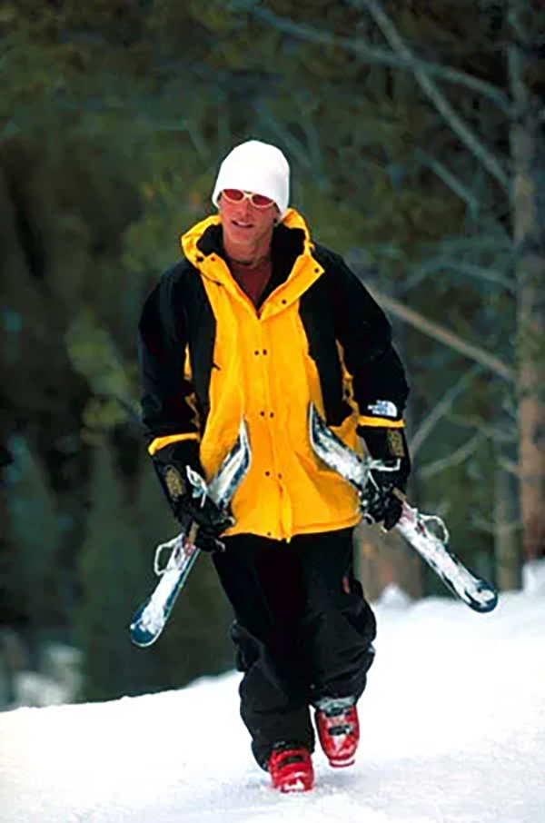 Man wearing a yellow jacket and sunglasses carrying ski blades on the snow